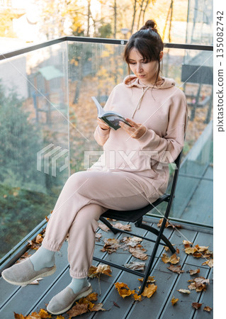 Woman holds open book on glass balcony during autumn. Intellectual wellness, self improvement, cognitive health, mindful reading. 135082742