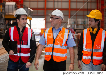 Three industrial engineers wearing safety uniform walk and discuss operations inside heavy factory, representing leadership, teamwork, and communication in high-risk industrial. Three industrial engineers wearing safety uniform walk and discuss operations inside heavy factory, representing leadership, teamwork, and communication in high-risk industrial. 135083054