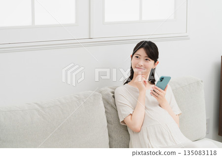 Pregnant woman operating a smartphone on the sofa Lifestyle image Pregnant woman operating a smartphone on the sofa Lifestyle image 135083118