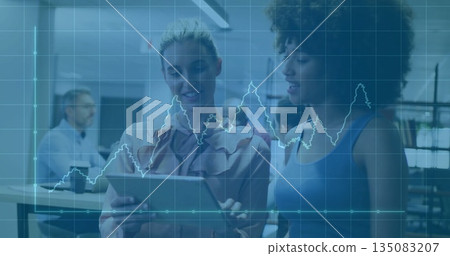 Two colleagues reviewing data on tablet in office, with coworker using laptop and chart overlay 135083207