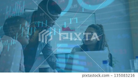 Colleagues analyzing financial charts around laptop and monitor in meeting room, with data overlays 135083216