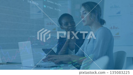 Female professionals in business attire collaborating in office, with laptop, tablet, data overlay 135083225