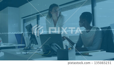 Discussing two colleagues leaning over desk toward laptop in office wearing business casual attire Discussing two colleagues leaning over desk toward laptop in office wearing business casual attire 135083231