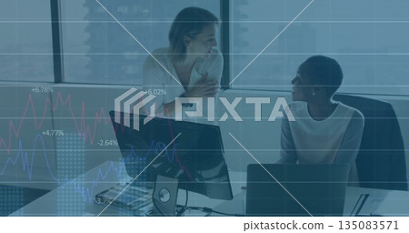 Two women in business attire discussing plans in office, with dual monitors laptop keyboard mouse 135083571