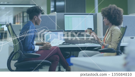 Businesswomen discussing report at desk in modern office, with laptop, dual monitors, coffee cup 135084810