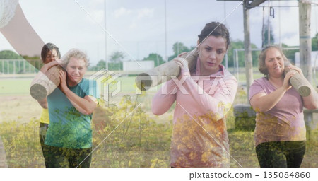 Striding women across grassy field carrying logs on shoulders, with metal rig and chain-link fence Striding women across grassy field carrying logs on shoulders, with metal rig and chain-link fence 135084860