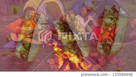 Strumming man in sweater playing guitar in forest glade, with campfire, lit tents, roasting skewers Strumming man in sweater playing guitar in forest glade, with campfire, lit tents, roasting skewers 135084896