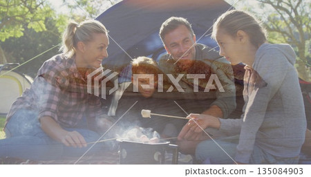 Family kneeling and sitting around camp stove roasting marshmallows in campsite, with dome tent 135084903