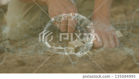 Hands carving soil with hand tool in dry sandy cracked earth outdoors, with wireframe grid overlay 135084995