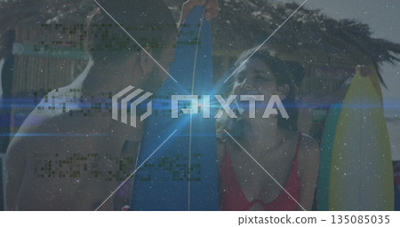 Laughing couple in swimwear holding surfboards in front of thatched-roof beach hut, palm fronds 135085035