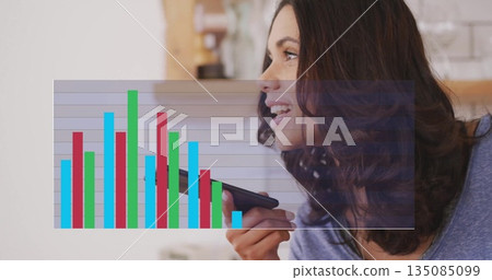 Speaking woman holding smartphone near mouth in home kitchen, with bar chart overlay, copy space 135085099
