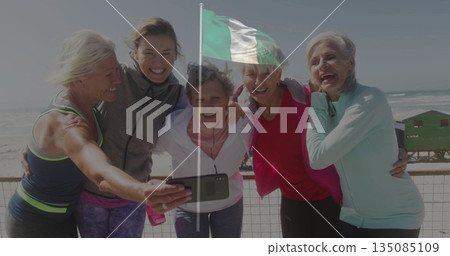 Smiling five women in workout clothes taking selfie on boardwalk, with smartphone railing and flag 135085109