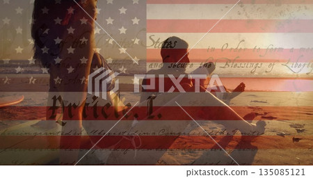 Relaxing couple in swimwear on sandy shore at sunset, with surfboard American flag script overlay 135085121