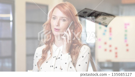 Talking red-haired woman with strap on phone, viewing dollar bill graphic in office with whiteboard Talking red-haired woman with strap on phone, viewing dollar bill graphic in office with whiteboard 135085127