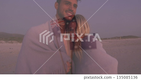 Standing couple leaning into each other on sandy beach at dusk, holding light-colored blanket 135085136