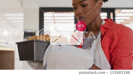 Baker wearing striped apron pulling metal pan with bread from oven in kitchen, with quilted mitts Baker wearing striped apron pulling metal pan with bread from oven in kitchen, with quilted mitts 135085199