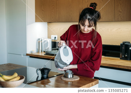 Person pours filtered water from pitcher into base of moka pot on wooden counter. Manual espresso routine, elevated home brewing, artisanal coffee gear, precision coffee making. Person pours filtered water from pitcher into base of moka pot on wooden counter. Manual espresso routine, elevated home brewing, artisanal coffee gear, precision coffee making. 135085271