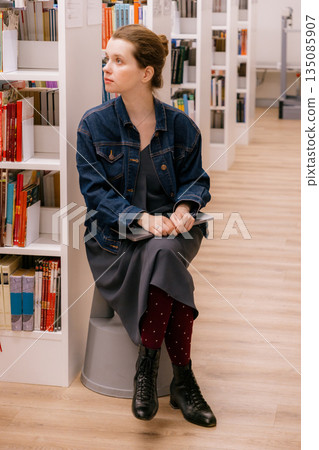 woman sits at shelves in library and reads book. education, scientific work. business person. 135085907