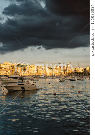 A picturesque view of Birgu Port filled with numerous boats, bathed in warm pre-sunset light. The tranquil waters reflect the golden hues, creating a serene maritime scene in Malta. 135086054