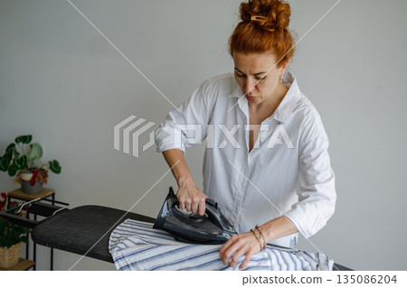 Young woman using ironing board to press clothes neatly at home. Young woman using ironing board to press clothes neatly at home. 135086204