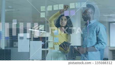 Placing sticky note, brainstorming plan in business attire on glass board in office, with tablet Placing sticky note, brainstorming plan in business attire on glass board in office, with tablet 135086312
