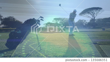 Swinging senior golfer wearing cap, hitting driver on tee box at golf course, with golf bag 135086351