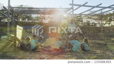 Teaching instructor holding paper and speaking to children under vine pergola, with campfire ring 135086365