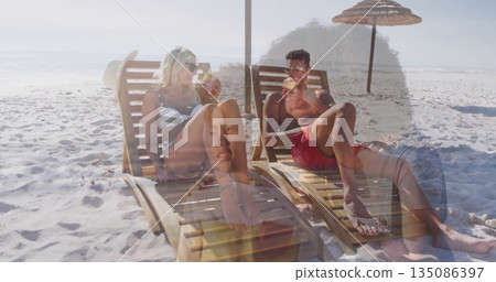 Couple wearing bikini and swim trunks sipping coconut drinks on beach, with chairs and umbrella 135086397