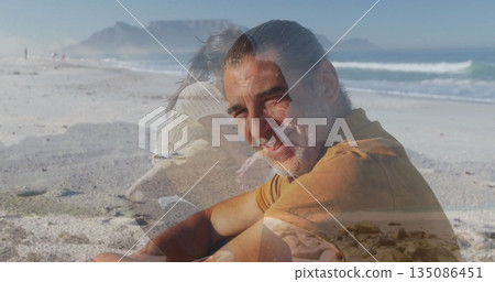 Smiling mature man wearing rust shirt and trousers sitting on sandy beach, with ocean waves 135086451