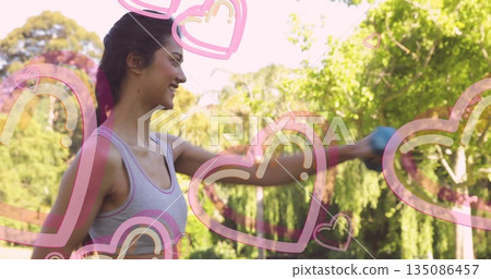 Standing sporty woman lifting blue dumbbell with floating pink heart graphics in garden, copy space Standing sporty woman lifting blue dumbbell with floating pink heart graphics in garden, copy space 135086457