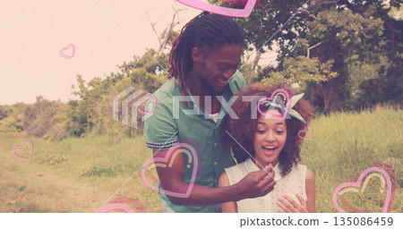Holding small object, couple standing in grassy field near woods, with translucent heart graphics 135086459