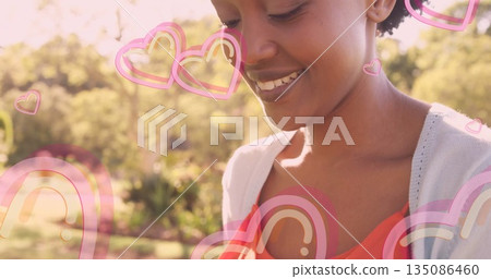 Smiling woman wearing red top gazing downward in sunlit park, with cartoon hearts, copy space 135086460