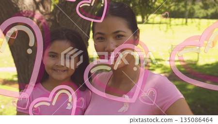 Smiling mother and daughter standing in park by tree trunk, with ribbon pins and heart overlays Smiling mother and daughter standing in park by tree trunk, with ribbon pins and heart overlays 135086464