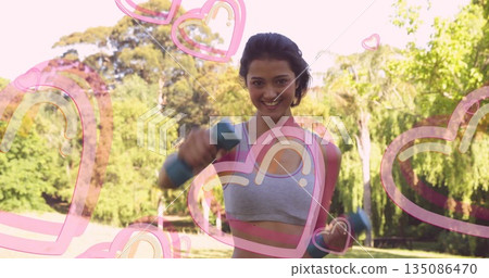 Holding two blue dumbbells, Asian woman in sportswear exercising in park, with pink heart overlays 135086470