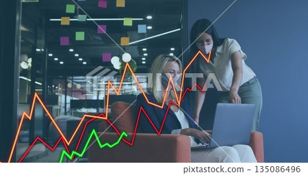 Collaborating women wearing face masks reviewing laptop line graph in lounge, with sticky notes 135086496