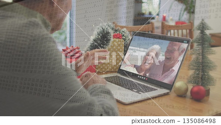 Senior man holding up gold-wrapped gift toward laptop in dining room, with pine tree baubles 135086498