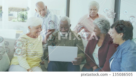 Gathering seniors wearing casual clothing collaborating around silver laptop in residential lounge Gathering seniors wearing casual clothing collaborating around silver laptop in residential lounge 135086505