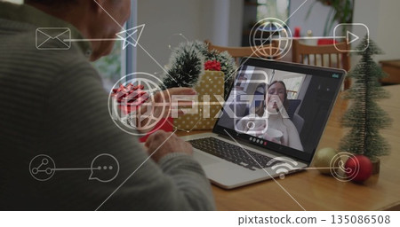 Talking senior male gesturing toward laptop during holiday call at home table, with trees and gifts 135086508
