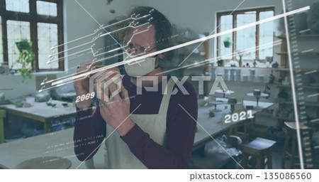 Inspecting masked artisan evaluating ceramic vessel in pottery studio, with digital data overlay 135086560