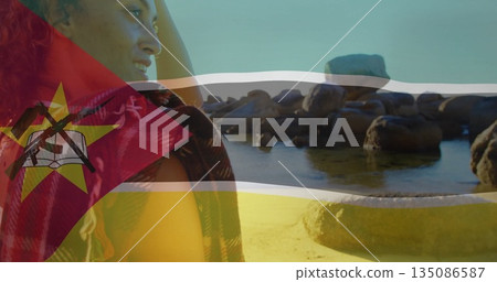 Standing woman wearing patterned shawl looking out at rocky beach, with Mozambique flag, copy space 135086587