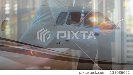 Holding smartphone and stylus, executive in suit leaning on jet wing in hangar, with tinted window Holding smartphone and stylus, executive in suit leaning on jet wing in hangar, with tinted window 135086632