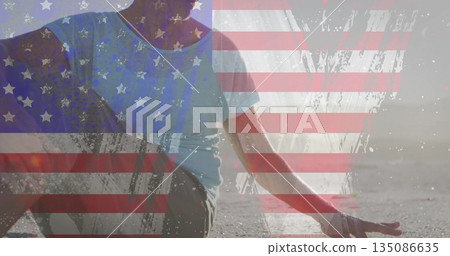 Sitting senior woman in T-shirt and leggings doing yoga on beach at sunset, with flag overlay 135086635