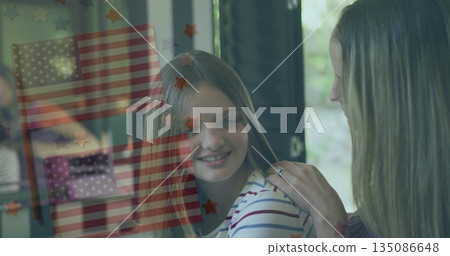 Mother adjusting hair of daughter wearing striped shirt near window with star decals, flag overlay 135086648