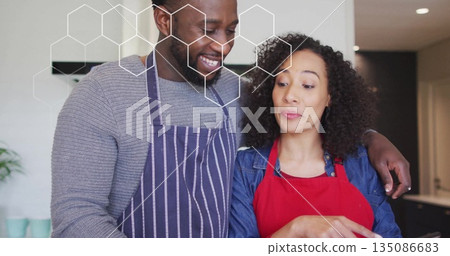Couple hugging and cooking in home kitchen, with striped apron, red apron and hexagonal overlay 135086683