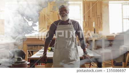 Standing African American carpenter wearing gray apron sanding wooden plank in carpentry workshop 135086697