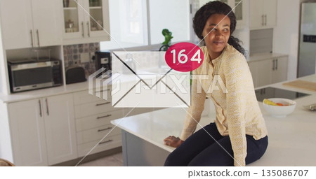 Glancing mid adult woman sitting on kitchen island at home, with envelope icon and fruit bowl Glancing mid adult woman sitting on kitchen island at home, with envelope icon and fruit bowl 135086707