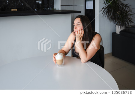 Cute, slightly sleepy young woman enjoying a warm cappuccino at home in a bright cozy kitchen. Simple morning routine, relaxed atmosphere, casual home lifestyle, calm start of the day. Cute, slightly sleepy young woman enjoying a warm cappuccino at home in a bright cozy kitchen. Simple morning routine, relaxed atmosphere, casual home lifestyle, calm start of the day. 135086758