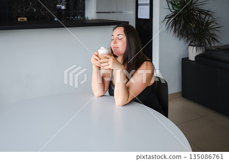 Cute, slightly sleepy young woman enjoying a warm cappuccino at home in a bright cozy kitchen. Simple morning routine, relaxed atmosphere, casual home lifestyle, calm start of the day. Cute, slightly sleepy young woman enjoying a warm cappuccino at home in a bright cozy kitchen. Simple morning routine, relaxed atmosphere, casual home lifestyle, calm start of the day. 135086761