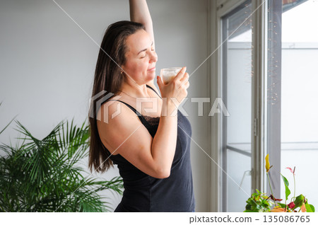 Cute, slightly sleepy young woman enjoying a warm cappuccino at home in a bright cozy kitchen. Simple morning routine, relaxed atmosphere, casual home lifestyle, calm start of the day. Cute, slightly sleepy young woman enjoying a warm cappuccino at home in a bright cozy kitchen. Simple morning routine, relaxed atmosphere, casual home lifestyle, calm start of the day. 135086765