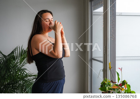 Cute, slightly sleepy young woman enjoying a warm cappuccino at home in a bright cozy kitchen. Simple morning routine, relaxed atmosphere, casual home lifestyle, calm start of the day. Cute, slightly sleepy young woman enjoying a warm cappuccino at home in a bright cozy kitchen. Simple morning routine, relaxed atmosphere, casual home lifestyle, calm start of the day. 135086768
